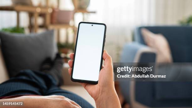 mujer usando el teléfono con pantalla blanca mientras está acostada en el sofá de su casa, pantalla de maqueta - mano agarrando fotografías e imágenes de stock