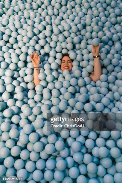 happy woman playing in a ball pool filled with plastic blue balls - adult ball pit stock pictures, royalty-free photos & images