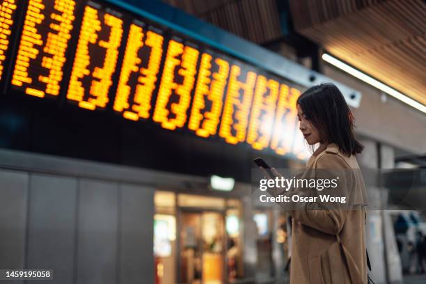 young asian woman using smartphone against arrival departure board at train station - estación de metro fotografías e imágenes de stock