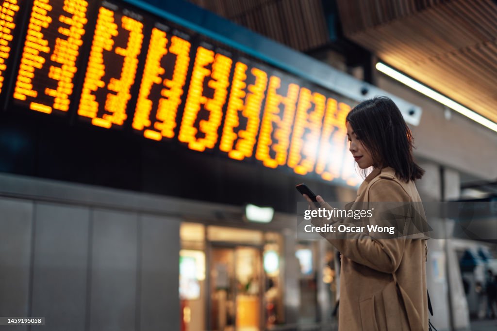 Young Asian woman using smartphone against arrival departure board at train station