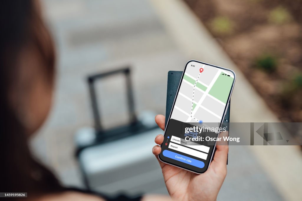Over the shoulder view of young woman using rideshare mobile app on smartphone to order taxi service