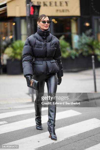Guest wears black sunglasses, a silver and diamonds Balenciaga logo earrings, a black high neck belted puffer jacket, black shiny leather legging...