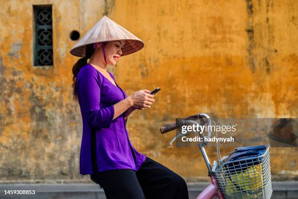vietnamese woman using mobile phone on a bicycle, old town in hoi an city, vietnam - traditional clothing stock pictures, royalty-free photos & images