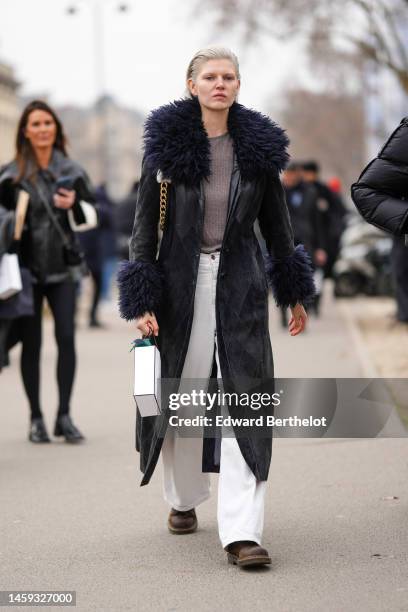 Guest wears a brown t-shirt, a black shiny leather with embroidered navy blue large fur collar long coat, white denim wide legs pants, a white...