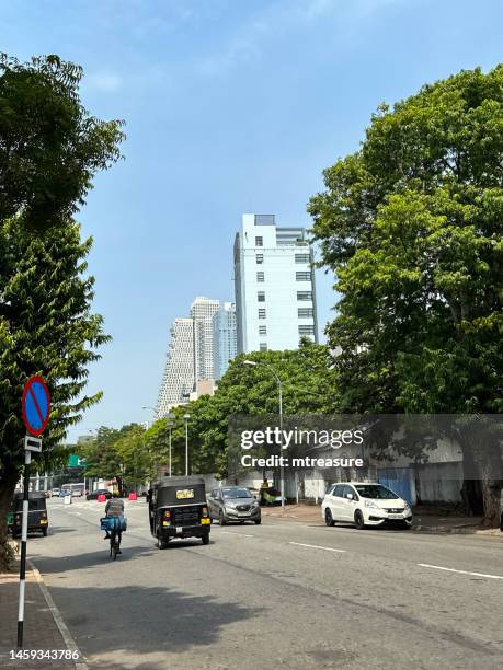 image of downtown colombo skyscrapers seen from neighbouring residential street, black and yellow auto rickshaw on treelined tarmac road, clear blue sunny sky background, focus on foreground - colombo stock pictures, royalty-free photos & images