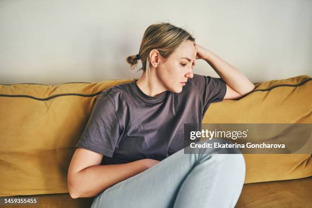 a young woman is sitting on the yellow couch at home with her head in her hands - despair stock pictures, royalty-free photos & images