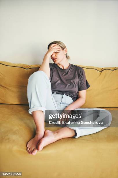 a young woman is sitting on the yellow couch at home with her head in her hands - stressed young woman sitting on couch stock-fotos und bilder