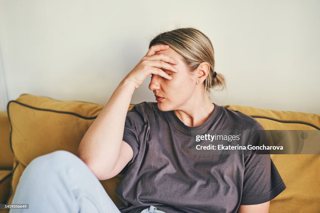 A young woman is sitting on the yellow couch at home with her head in her hands
