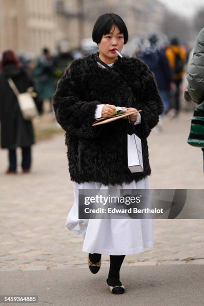 Guest wears a white long dress, black tights, a white shopping bag from Chanel, a black oversized fur, beige leather and black toe-cap ballerinas ,...