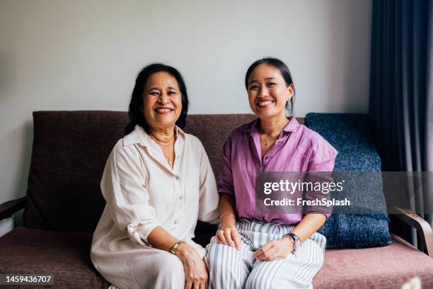 a happy beautiful senior woman and her pretty daughter looking at camera while sitting on the cozy sofa - cozy home stock pictures, royalty-free photos & images