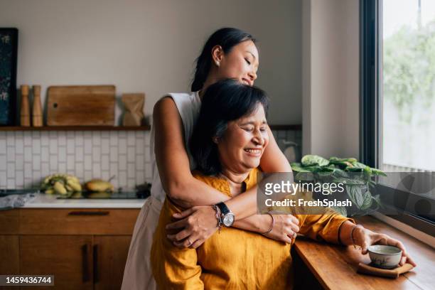 eine glückliche schöne frau, die ihre mutter umarmt, während sie in der küche sitzt und tee trinkt - beautiful granny stock-fotos und bilder