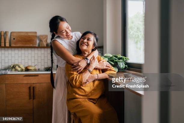 a happy beautiful woman hugging her mother while she is sitting in the kitchen and drinking tea - asia stock pictures, royalty-free photos & images