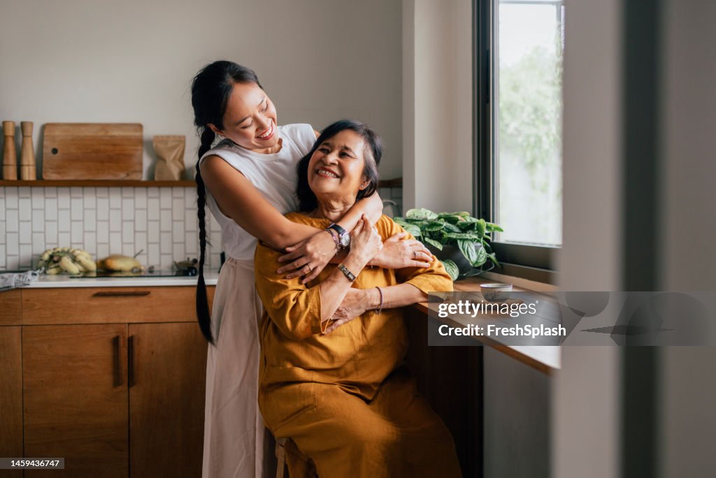 A Happy Beautiful Woman Hugging Her Mother While She Is Sitting In The Kitchen And Drinking Tea