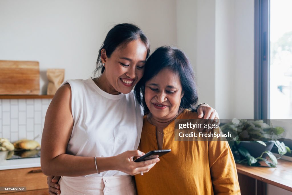 A Happy Beautiful Woman Watching Something On Her Mobile Phone With Her Mother (She Is Embracing Her) While They Are Standing In The Kitchen