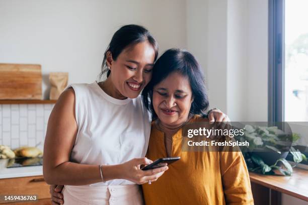 a happy beautiful woman watching something on her mobile phone with her mother (she is embracing her) while they are standing in the kitchen - asian mother stock pictures, royalty-free photos & images