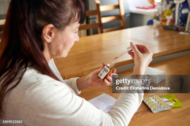 adult woman holds a bottle of sbd oil with a pipette in her hands while sitting kitchen table - mature woman herbs stock pictures, royalty-free photos & images