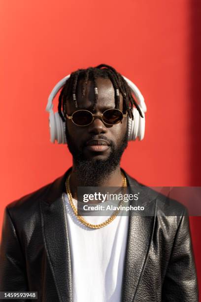 portrait of black man looking at camera while listening to music with red background on a sunny day - vestimenta stock pictures, royalty-free photos & images