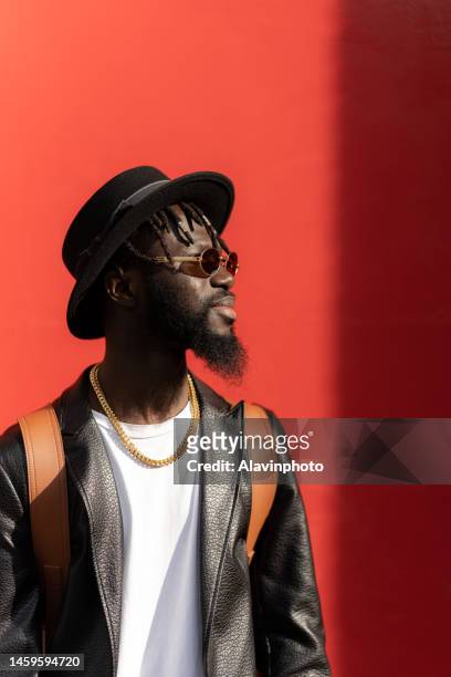 portrait of black man looking into camera with red background on a sunny day - vestimenta stockfoto's en -beelden