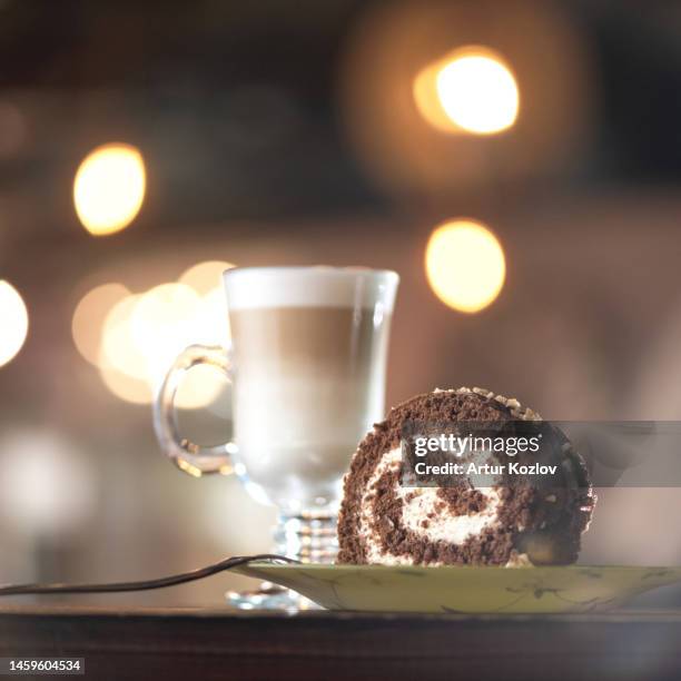 coffee latte with portion of chocolate roll on table. coffee coffee in glass cup and portioned piece of roll on plate with fork. serving dish. background of blurry lights - soirée mousse stock-fotos und bilder