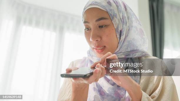 nahaufnahme der glücklichen jungen muslimischen frau sitzt auf dem bett mit computer-laptop und verwenden smartphone sprachbefehle an virtuelle assistenten im schlafzimmer zu hause. arbeiten sie von zu hause aus. - übersetzung stock-fotos und bilder
