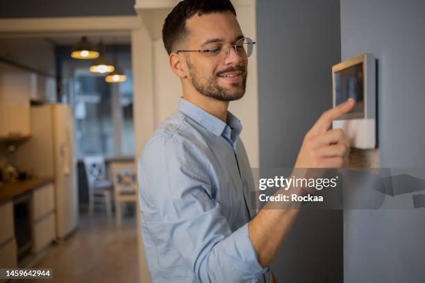 un jeune homme ajuste la température à la maison avec un appareil fixé au mur - thermostat photos et images de collection