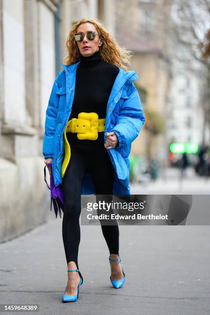 Guest wears black large sunglasses, a black turtleneck pullover, black legging pants, a blue oversized puffer jacket, a yellow shiny leather large...