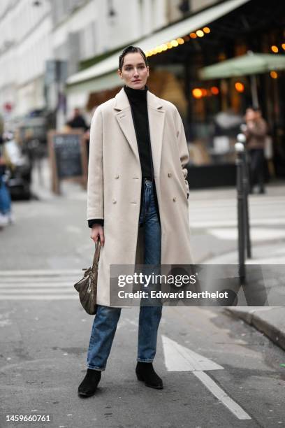 Guest wears a black turtleneck wool pullover, a beige long coat, blue denim cigarette pants, black suede pointed ankle boots , a gold shiny handbag ,...