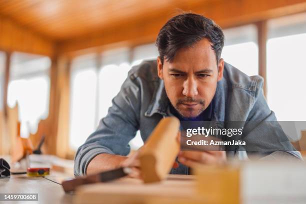 frontal view of a young dark-skinned man holding and observing carefully a piece of wood that he is working on - human face frontal stock pictures, royalty-free photos & images