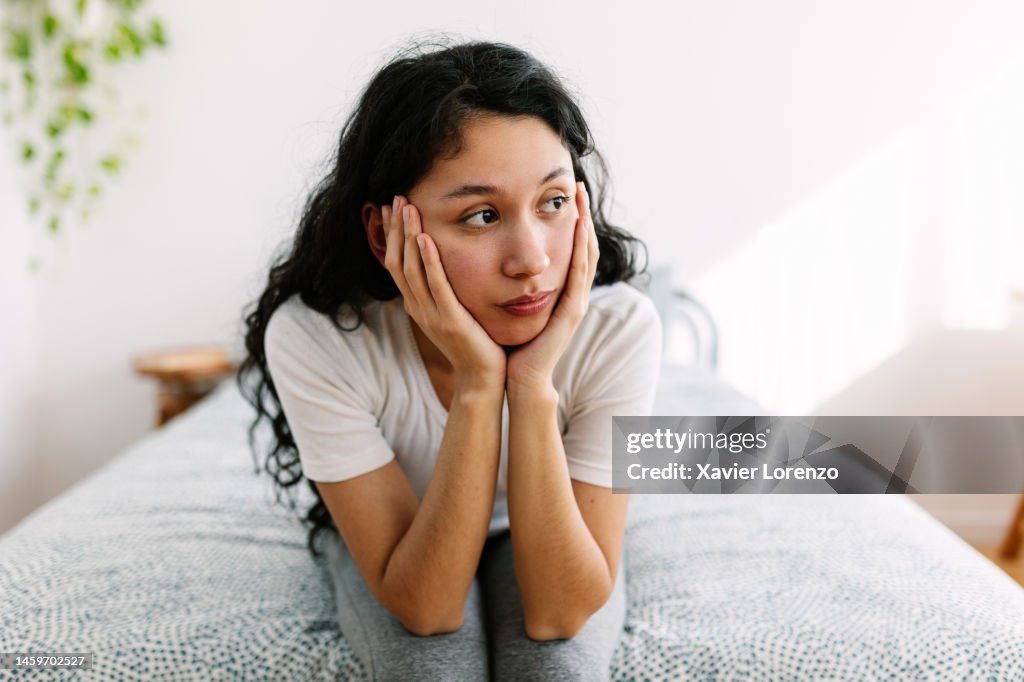 Sad millennial woman sitting on bed looking away thinking about problems and bad feelings