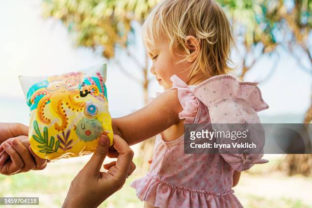 mother putting on kid's swimming floats ring arm sleeve or arm floats for her little daughter  beautiful baby girl in a swimsuit during family vacation along beach. - beach babes stock pictures, royalty-free photos & images