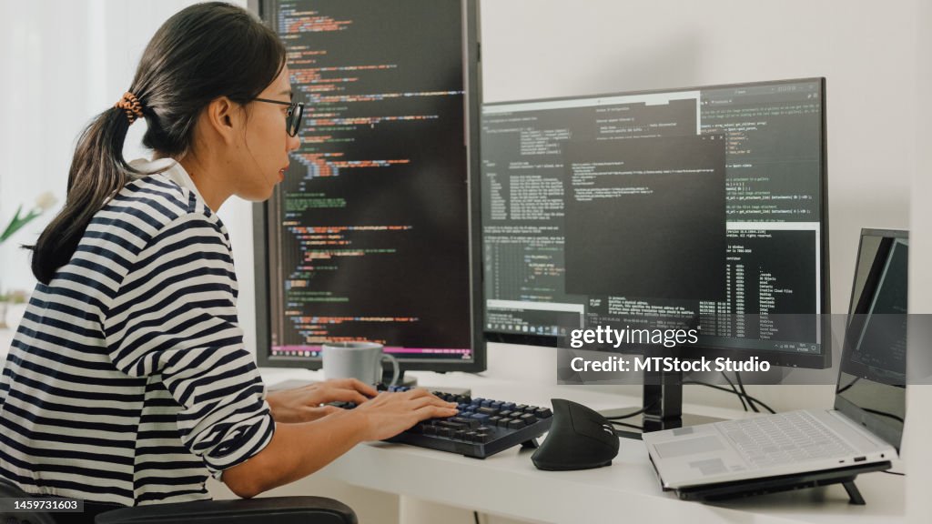 Young Asian woman software developers using computer to write code sitting at desk with multiple screens work remotely at home.