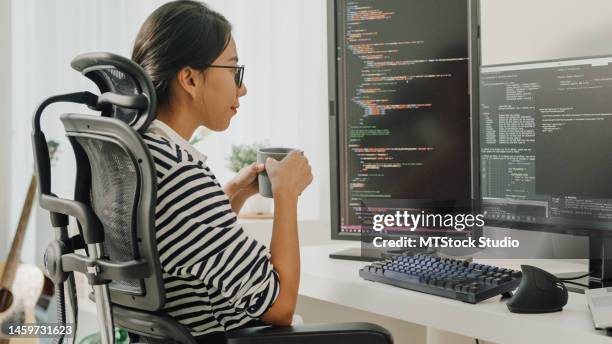 junge asiatische softwareentwicklerinnen, die kaffee trinken und computer verwenden, um code zu schreiben, sitzen am schreibtisch mit mehreren bildschirmen und arbeiten remote zu hause. - html stock-fotos und bilder