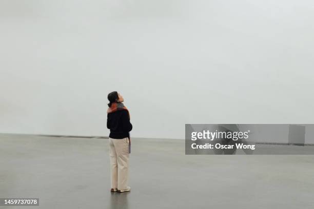 full length of young asian woman looking up while standing against white wall - exposition wall ストックフォトと画像