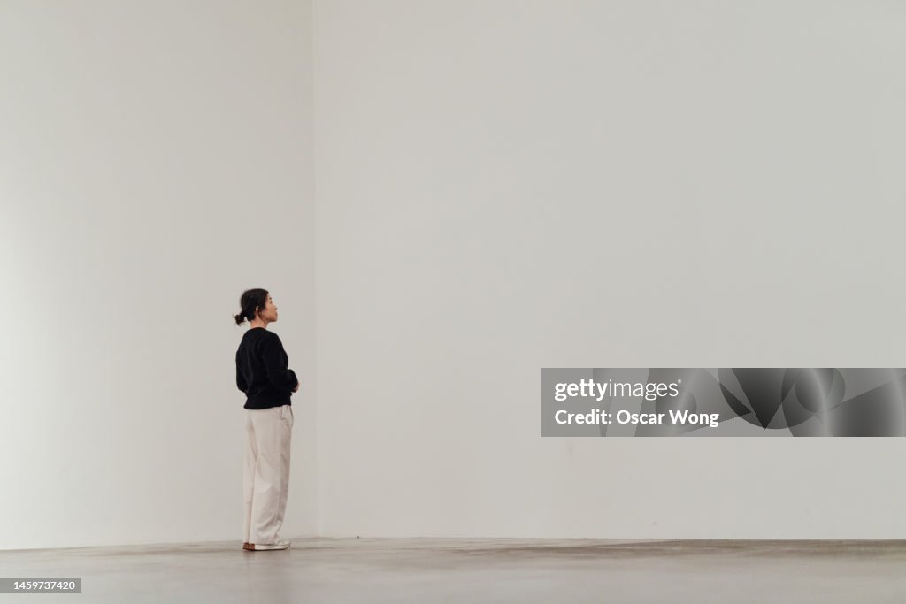 Full length of young Asian woman looking away while standing against white wall