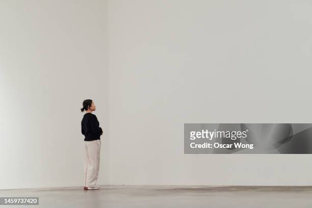 full length of young asian woman looking away while standing against white wall - stehen stock-fotos und bilder
