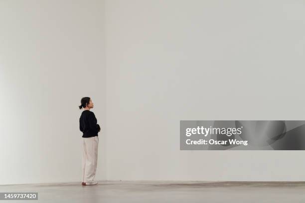 full length of young asian woman looking away while standing against white wall - art gallery people fotografías e imágenes de stock