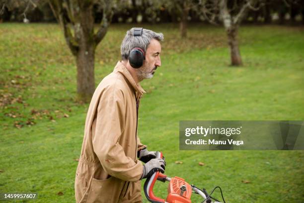 gardener in uniform and with ear protector cutting grass with lawn mower in a garden. - man mowing stock pictures, royalty-free photos & images