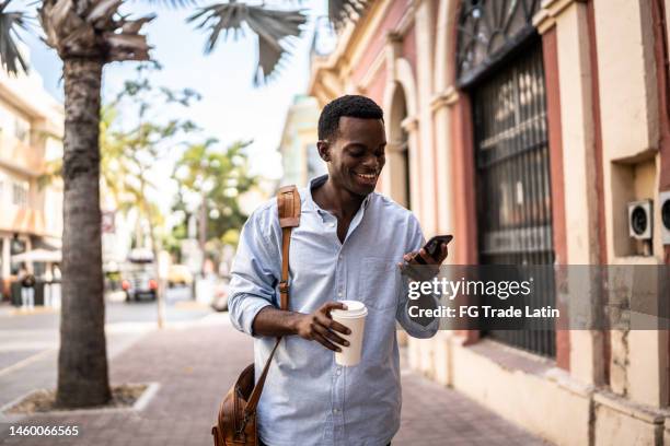 young man using cellphone while walking outdoors - messenger bag stock pictures, royalty-free photos & images
