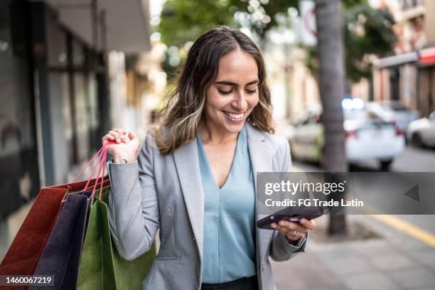 mid adult businesswoman using phone and carrying shopping bags outdoors - latin american and hispanic shopping bags stock pictures, royalty-free photos & images