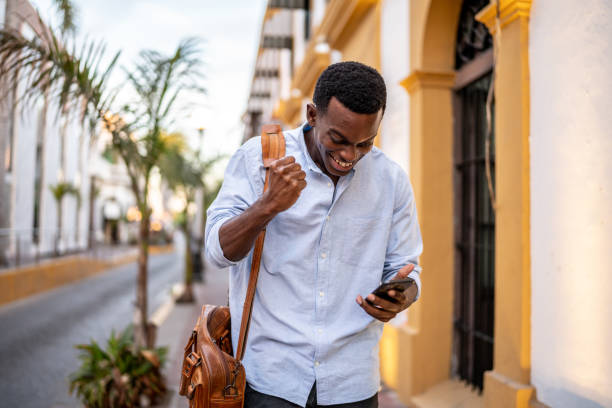 young man celebrating using mobile phone while walking outdoors - black person using phone celebrating stock pictures, royalty-free photos & images
