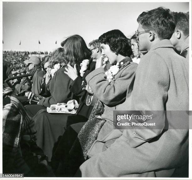 View of spectators, including a young woman wearing a corsage and eating a hot dog, in the stands at a college football game , East Lansing,...