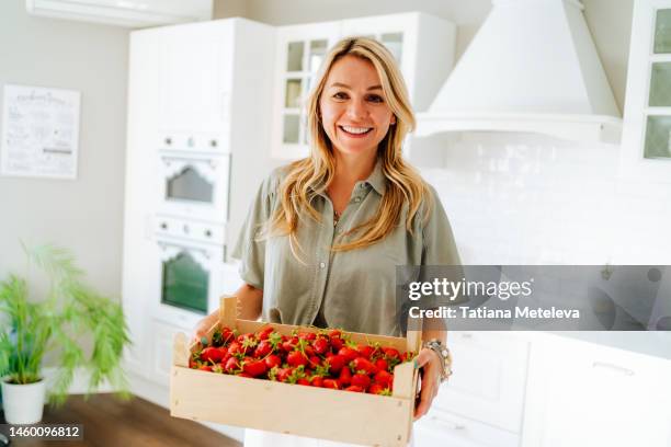 fresh and raw berry ingredient for any meal, season of harvest. blonde woman carrying strawberry box in hands in the white kitchen - fruit carton stock pictures, royalty-free photos & images