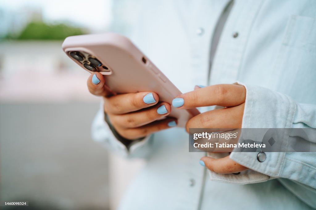 Close up female hands with blue manicure using pink smartphone outdoors