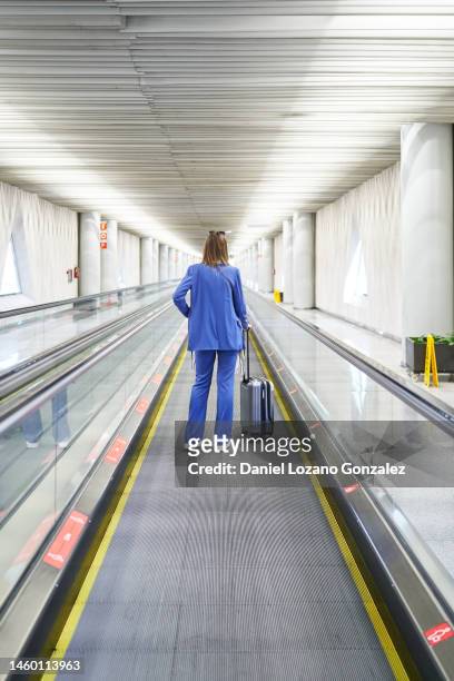 back view of unrecognizable woman standing in the airport moving walkway with her travel suitcase. - travolator stock pictures, royalty-free photos & images