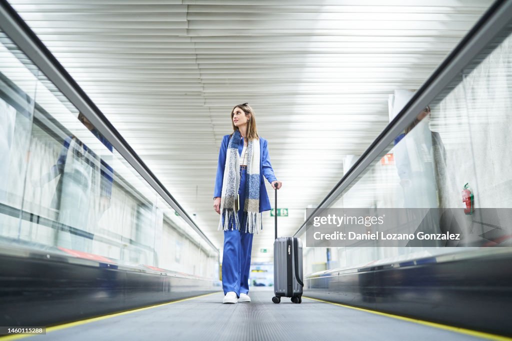 Cheerful woman standing in the airport moving walkway with her travel suitcase and looking away.