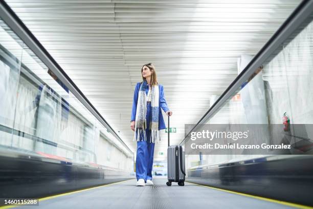 cheerful woman standing in the airport moving walkway with her travel suitcase and looking away. - travolator stock pictures, royalty-free photos & images