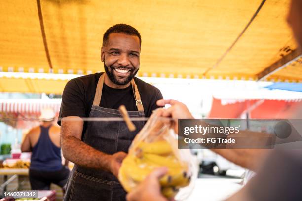 vendedor ayudando a su cliente a poner los plátanos en una bolsa de plástico en un mercado callejero - puesto de mercado fotografías e imágenes de stock
