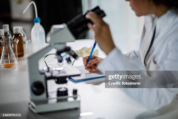 ripresa di una scienziata che scrive note su un blocco appunti dopo aver analizzato campioni al microscopio - epidemiologia foto e immagini stock