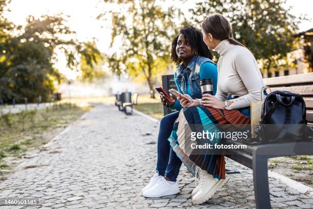 portrait of two friends sitting on the street bench, using smart phones and casually chatting over coffee - girlfriend meme stock pictures, royalty-free photos & images