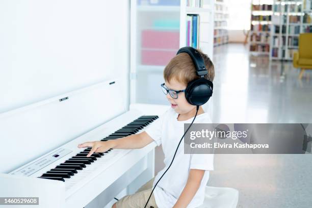 a little boy in glasses, big headphones and a white t-shirt plays a white digital piano in a music class - child pianist stock pictures, royalty-free photos & images